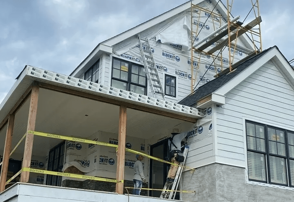 image (2) A house under construction with scaffolding, workers, and exterior walls partially covered in building wrap. Yellow caution tape marks off the porch area.