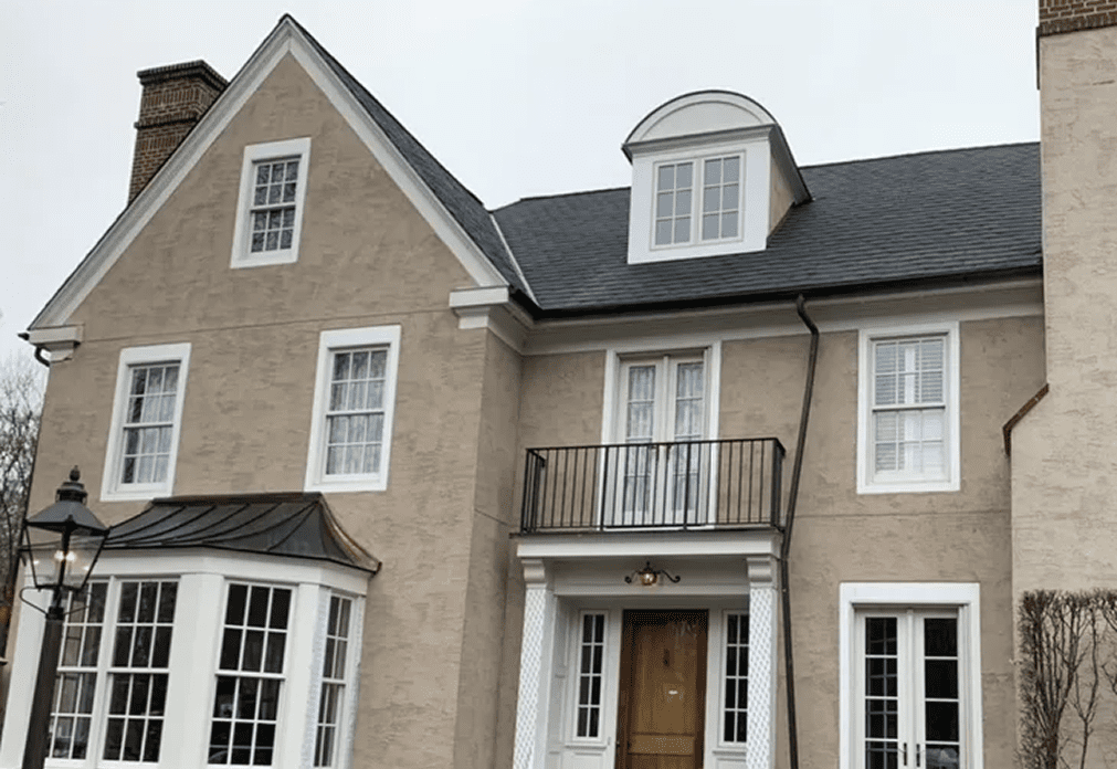 image A two-story beige house with white trim, black shingle roof, central wooden door, balcony above the entrance, and multiple windows on a cloudy day.