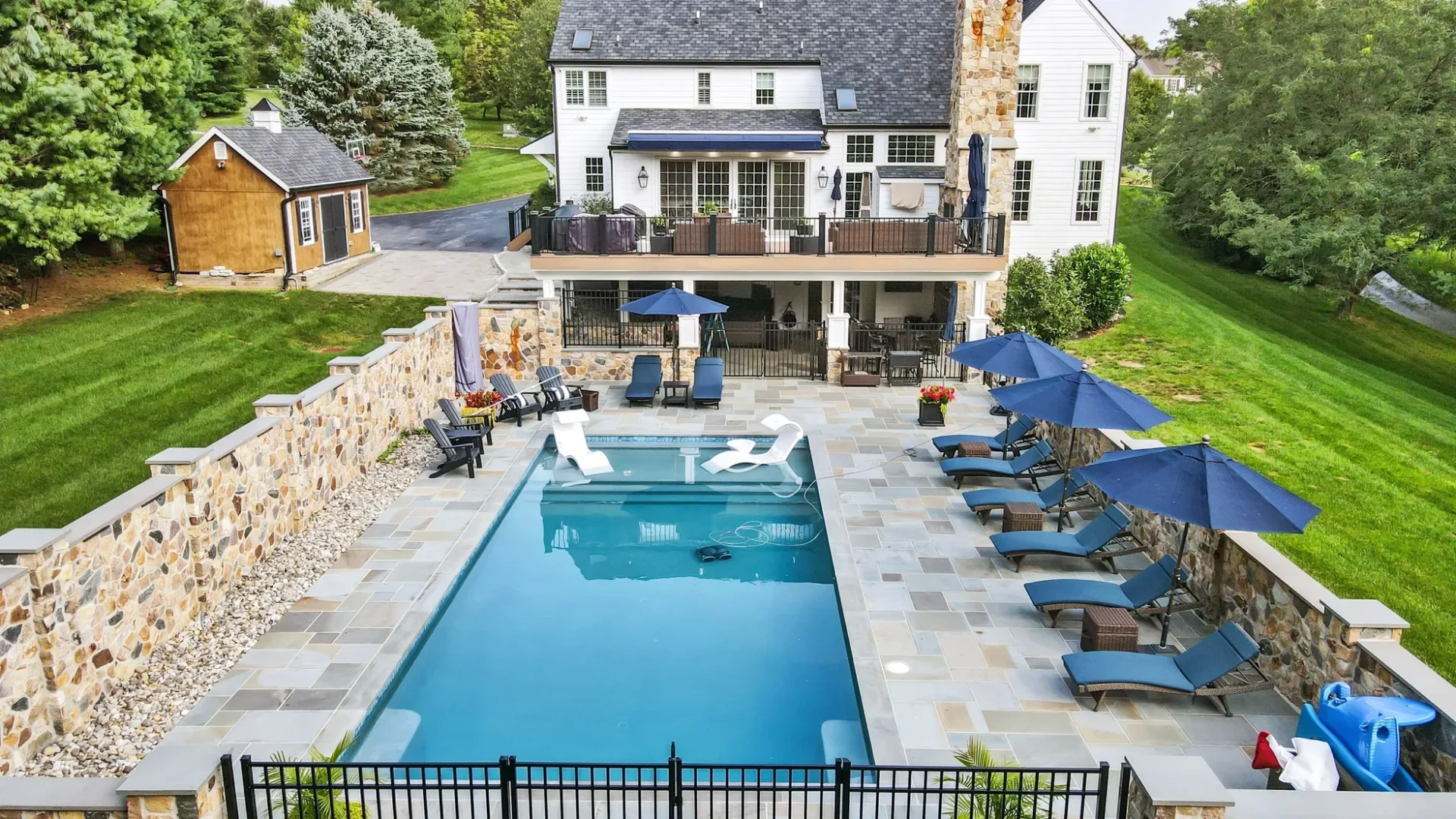 A rectangular backyard pool with lounge chairs, umbrellas, and two pool floats, bordered by stone walls and a large white house in the background.