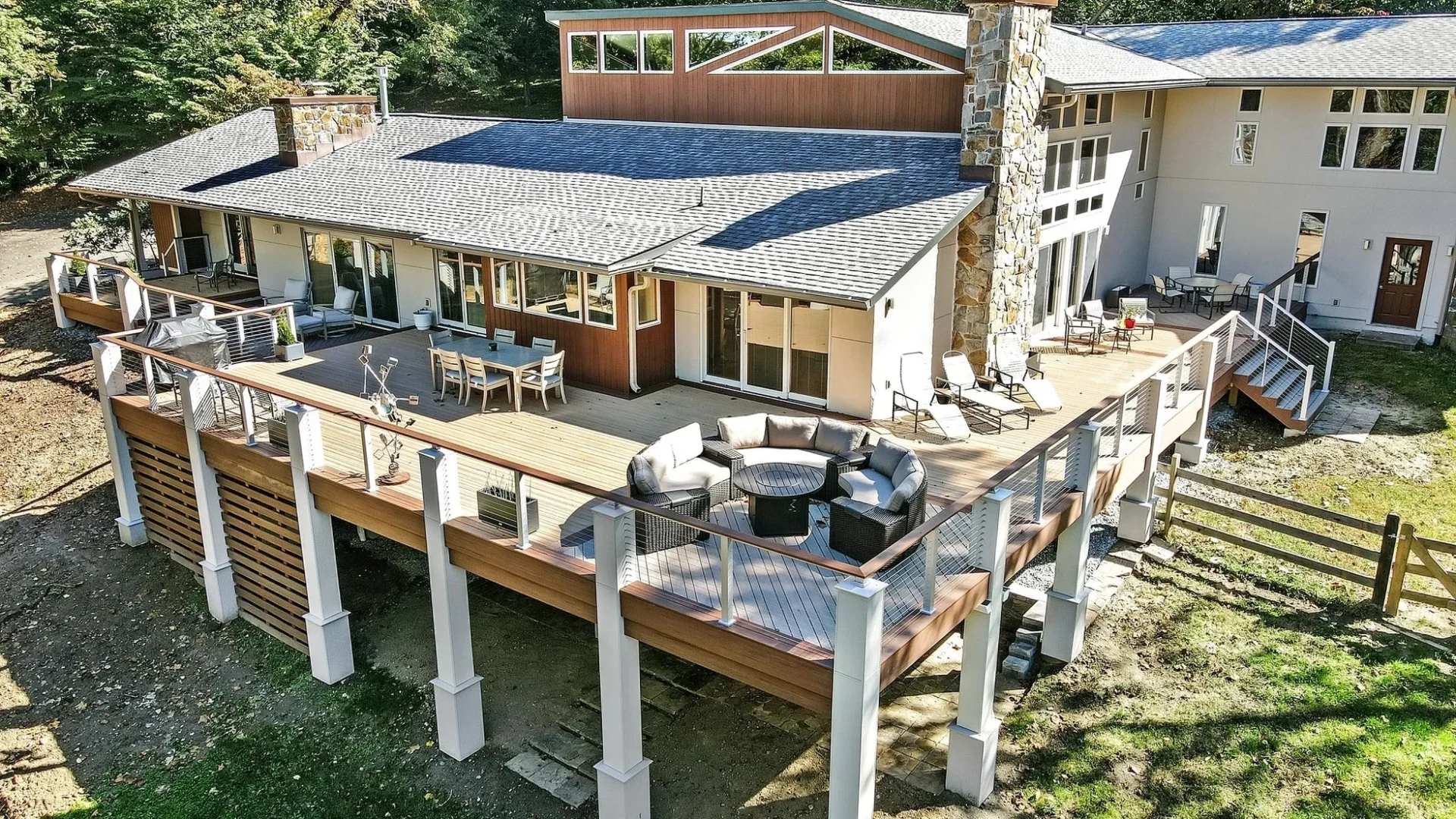 A modern house with large windows and a spacious elevated wooden deck featuring outdoor furniture and glass railing, surrounded by trees.
