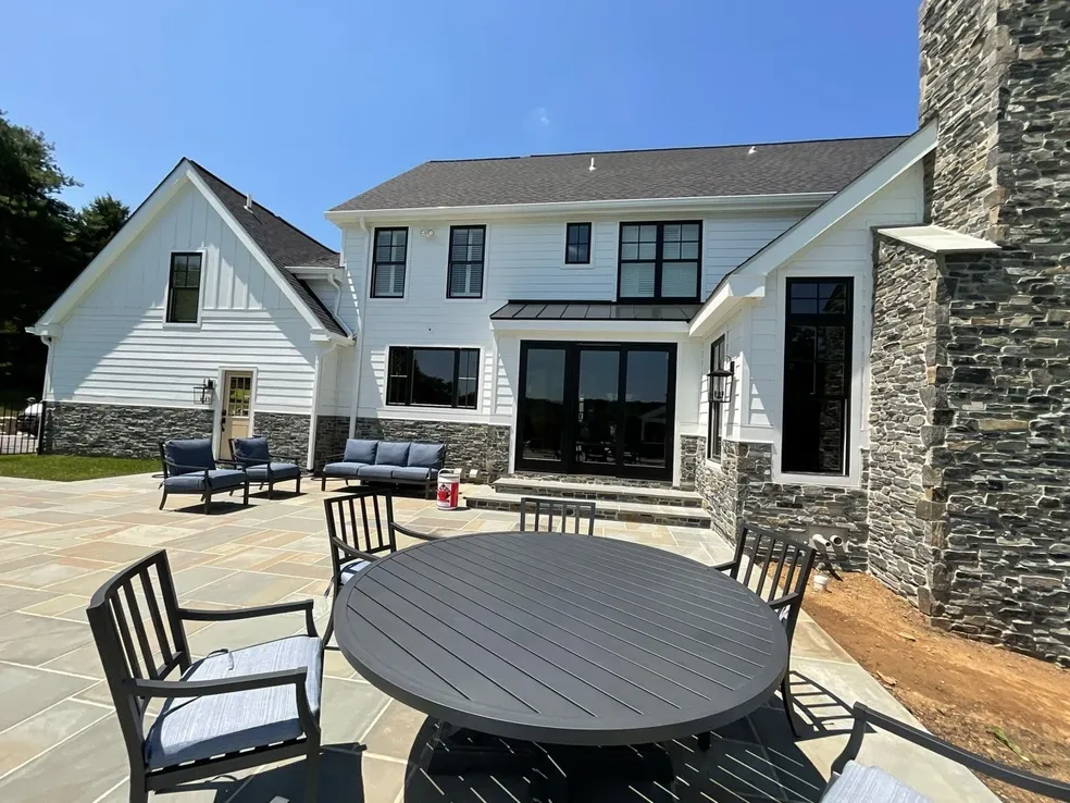 A modern house with white siding and stone accents features a large patio with outdoor seating and a round table under clear, sunny skies.