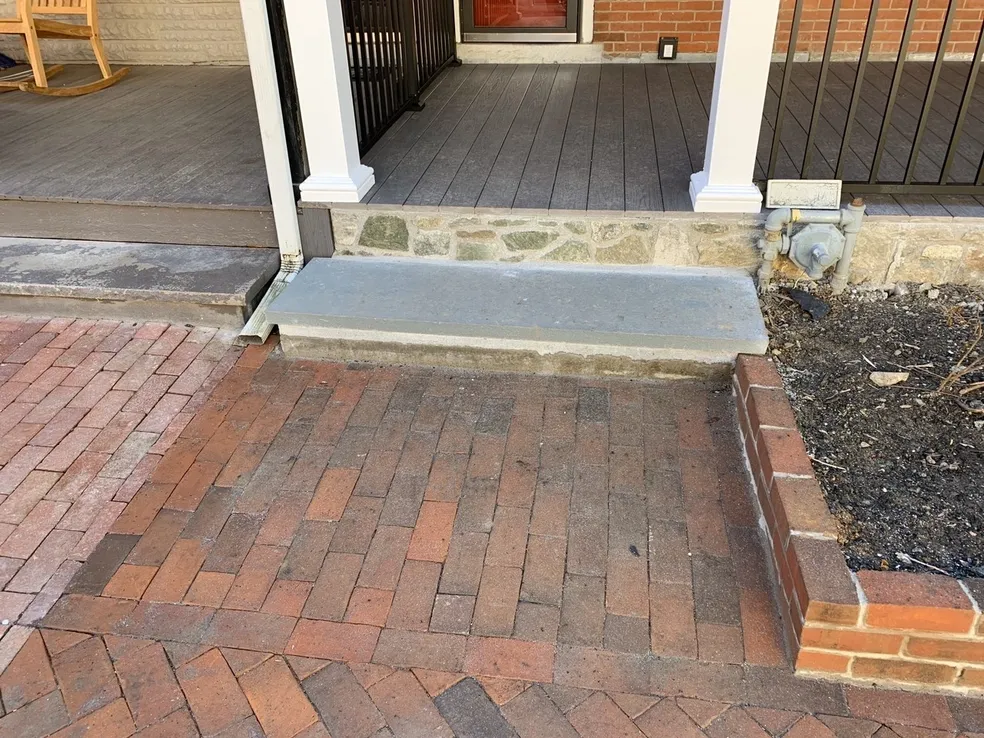 A concrete step leads up from a brick walkway to a porch with gray decking, bordered by a small stone wall and a raised garden bed.