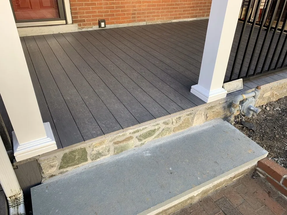 A raised porch with gray composite decking, white columns, and a stone step below, bordered by brick and a garden bed.