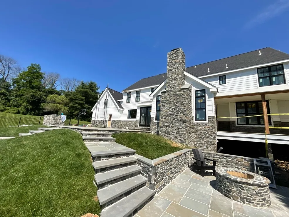 A modern house with stone accents, large windows, and a prominent outdoor stone staircase leading to a patio with a fire pit under a clear blue sky.