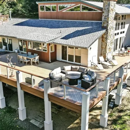 rs=w_800,cg_true (2) A modern house with large windows and an L-shaped elevated wooden deck featuring outdoor seating areas and surrounded by trees.